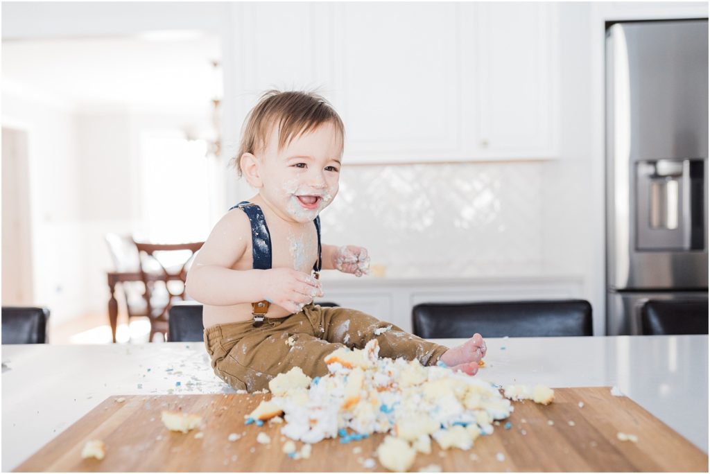 One year old boy first birthday cake smash photo session at home in New Jersey with a family of four in their beautiful white and grey kitchen. Sitting on the kitchen counter top eating cake. 
NJ Family Photographers Renee Ash Photography 