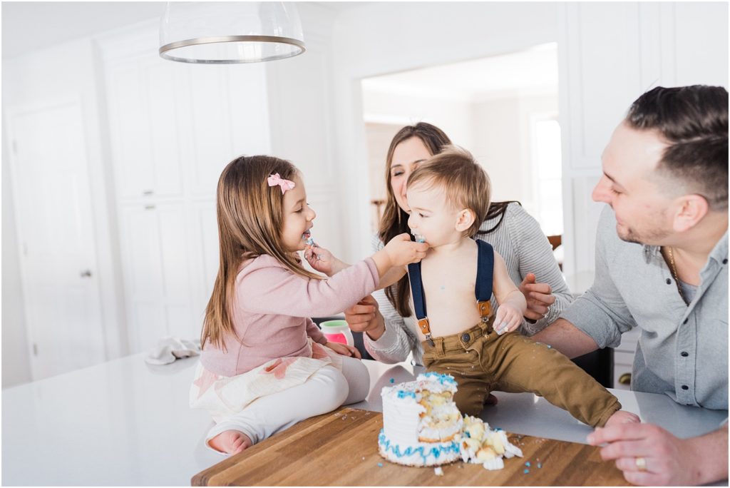 cake smash at home in New Jersey with a family of four in their beautiful white and grey kitchen. Sitting on the kitchen counter top eating cake. 
NJ Family Photographers Renee Ash Photography 