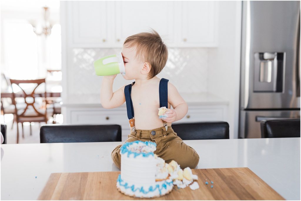 One year old boy first birthday cake smash photo session at home in New Jersey with a family of four in their beautiful white and grey kitchen. Sitting on the kitchen counter top eating cake. Drinking from a Munchkin Miracle 360 sippy cup
NJ Family Photographers Renee Ash Photography 