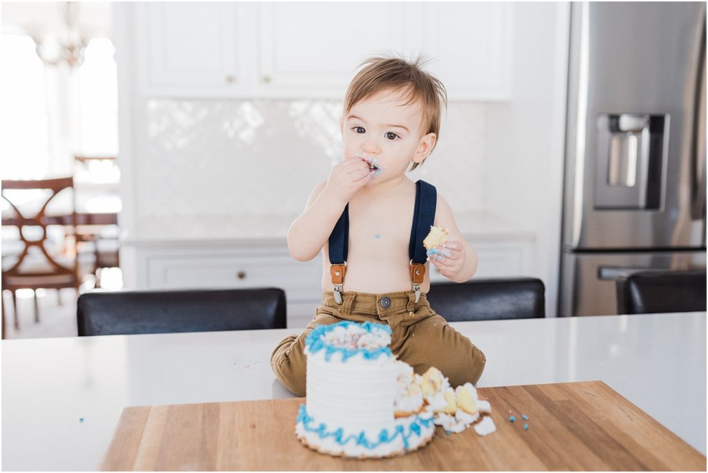 One year old boy first birthday cake smash photo session at home in New Jersey with a family of four in their beautiful white and grey kitchen. Sitting on the kitchen counter top eating cake. 
NJ Family Photographers Renee Ash Photography 