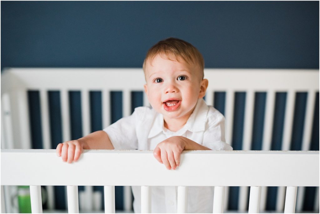  One year old boy in a white shirt standing in a white crib with navy blue walls. lifestyle First birthday pictures at home. by Renee Ash New Jersey photographers