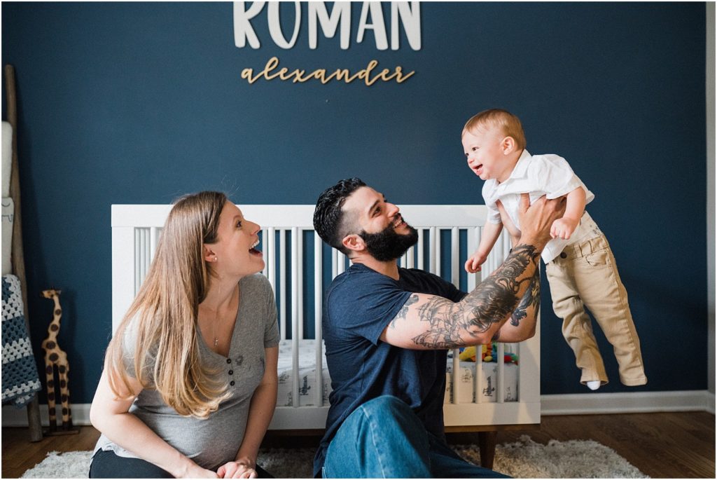 Family of three with their one year old boy in a white shirt sitting against a white crib in a navy blue, white, and gold nursery. lifestyle First birthday pictures at home. by Renee Ash New Jersey photographers