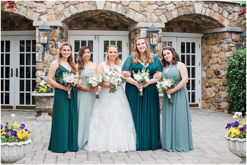 Bride and her four bridesmaids in mix and match green bridesmaid gowns and white and green bouquets by Olivia floral designs. Bridal Party photos in the courtyard at The Olde Mill Inn Wedding Venue in Northern NJ. by Renee Ash Photography 
