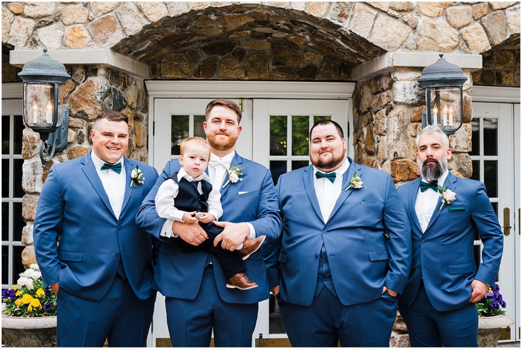 three Groomsmen and the groom in Navy blue suits and green bow ties. holding a toddler Ring bearer. Spring at The Olde Mill Inn Wedding Venue in Northern NJ. by Renee Ash Photography 