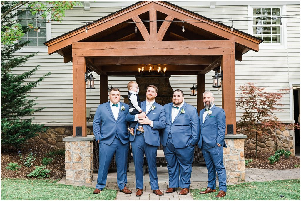 three Groomsmen and the groom in Navy blue suits and green bow ties. holding a toddler Ring bearer. Spring at The Olde Mill Inn Wedding Venue in Northern NJ. by Renee Ash Photography 