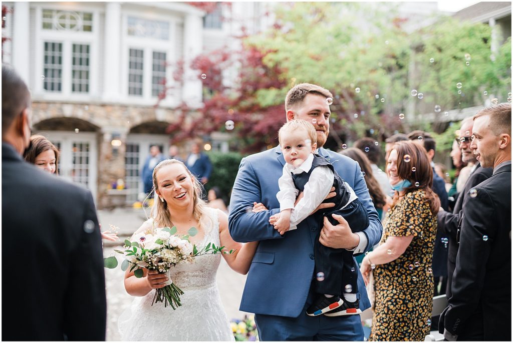 The bride and Groom carrying their toddler son back down the aisle as a family of three and husband and wife.  Bubbles blowing by guests after the first kiss In the courtyard ceremony space of The Olde Mill Inn Wedding Venue in Northern NJ. by Renee Ash Photography 