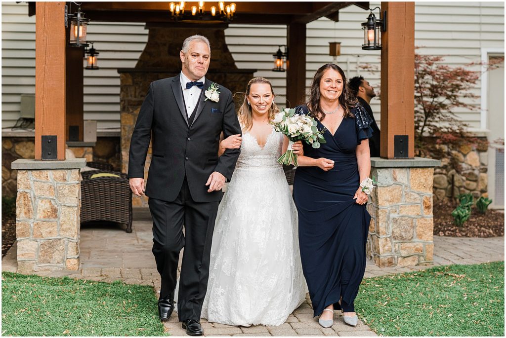 Bride being walked down the aisle by her mom and dad in the courtyard ceremony space of The Olde Mill Inn Wedding Venue in Northern NJ. by Renee Ash Photography 