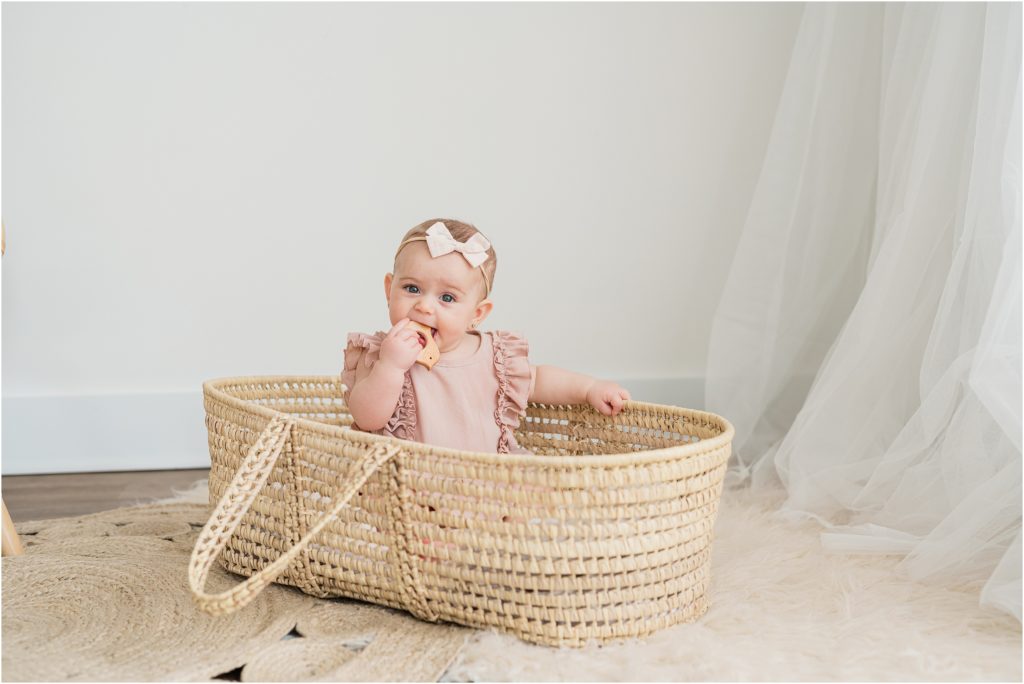 Six month old baby pictures in a woven baby bassinet wearing a mauve romper by Bailey's blossoms. Boho baby pictures in the Renee Ash Photography photo studio at the Shoppes at Lafayette, Lafayette NJ