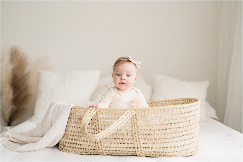 six month old baby girl on a white bed in a wicker baby moses basket. Wearing a neutral romper and ivory baby hair bow. The Shoppes at Lafayette photo studio. 