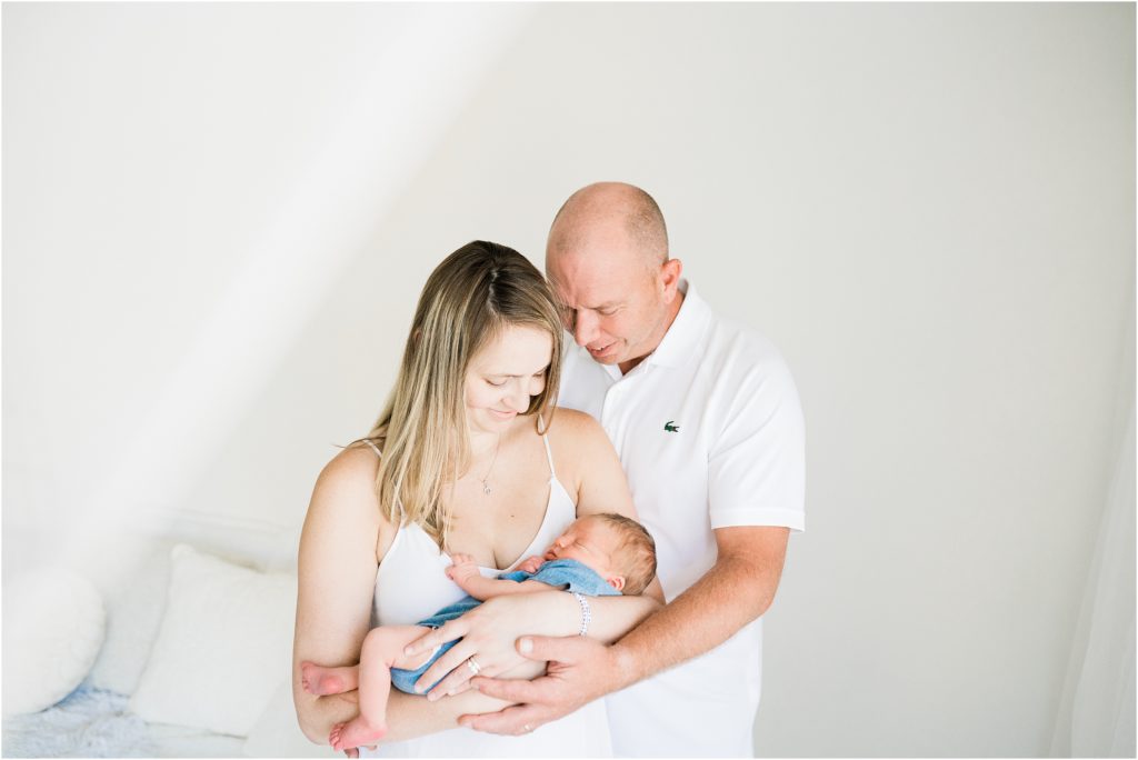planning a new jersey newborn session with Mom and dad holding a baby boy wrapped in blue in an all white studio. Renee Ash Photography, Sussex County NJ photo studio